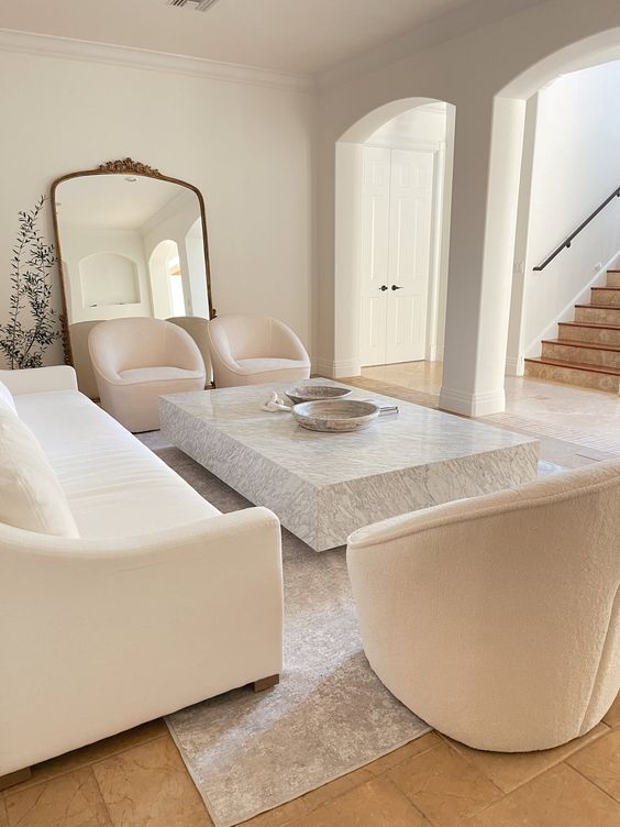 a beautiful white living room with a white sofa and creamy chairs, a low stone slab coffee table and a large floor mirror