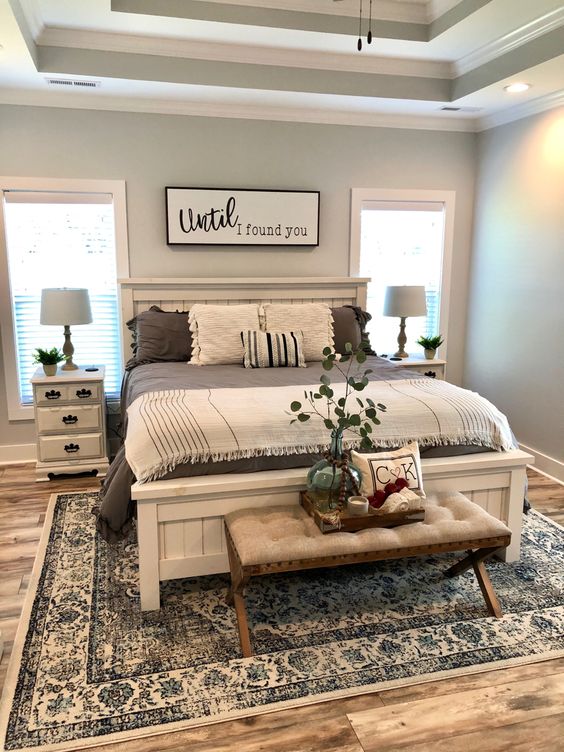 a blue farmhouse bedroom with a whitewashed bed, an upholstered bench, whitewashed nightstands, printed bedding and lights
