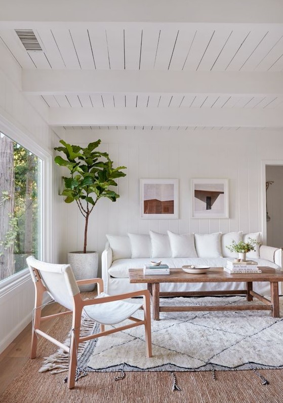 a boho living room with white furniture, a wooden coffee table, a small gallery wall and a tree