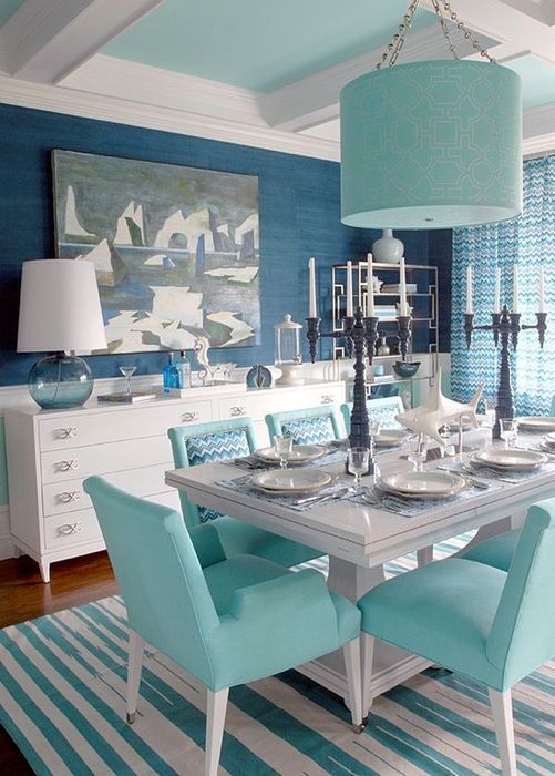 a bold dining room with navy walls and white paneling, white sideboards, a white dining table and turquoise chairs and a pendant lamp