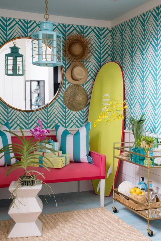 a bold tropical space with turquoise printed wallpaper, a pink bench with striped pillows, a round mirror, a gold cart and a turquoise pendant lamp