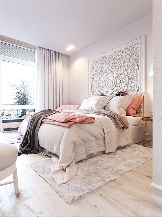 a chic bedroom with a whitewashed carved headboard, a bed with neutral bedding, a fluffy rug, white chairs and a large window