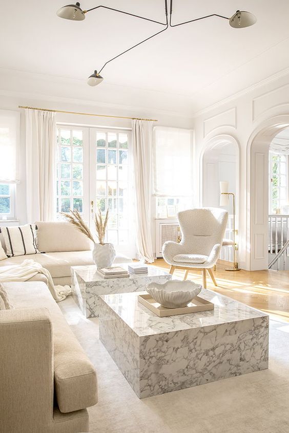 a chic white living room with a sectional, a duo of stone slab coffee tables, a white chair, white textiles and a chic chandelier