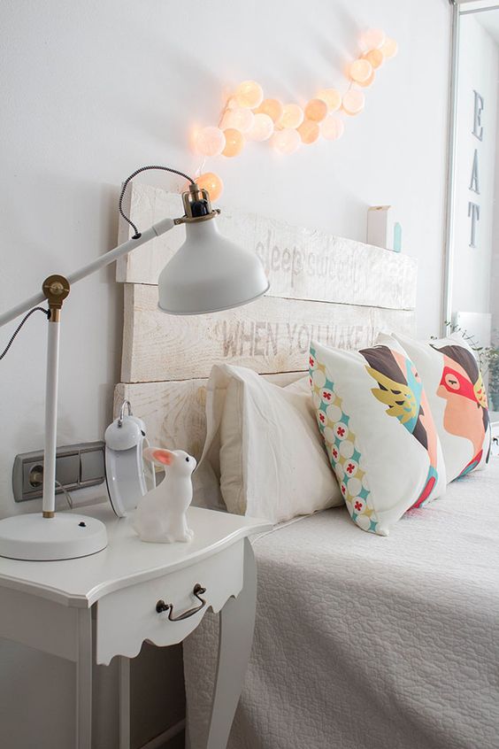 a cute kid bedroom in white, with a whitewashed planked headboard, lights over the bed, a white vintage nightstand and bright pillows