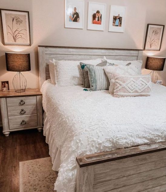 a farmhouse bedroom with a whitewashed bed and nightstands, table lamps, a gallery wall, neutral and pastel bedding, a fluffy rug
