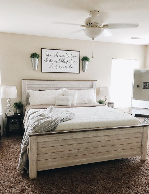 a farmhouse bedroom with tan walls, a whitewashed planked bed with neutral bedding, black nightstands, table lamps and buckets with greenery
