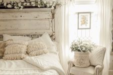 a lovely and welcoming white bedroom with a bed and a whitewashed headboard, a crystal chandelier, a creamy chair and a jute rug