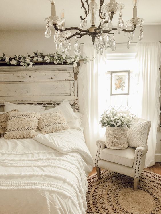 a neutral farmhouse bedroom with a whitewashed bed and an extensive headboard, a white chair, a crystal chandelier, a jute rug, neutral bedding