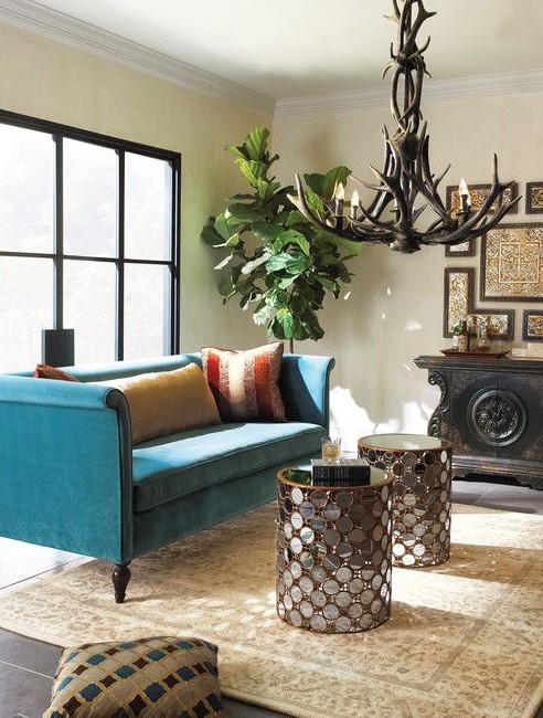 a refined living room with a glazed wall, a turquoise sofa, glass clad coffee tables, an antler chandelier, a refined gallery wall and a statement plant