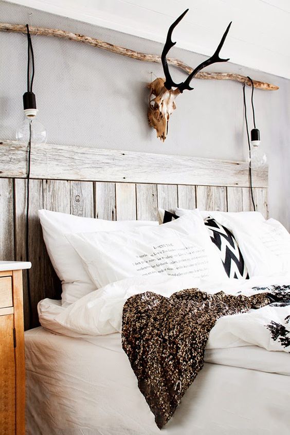 a rustic bedroom with a bed with a whitewashed and weathered headboard, white and printed bedding, a branch with bulbs and a skull