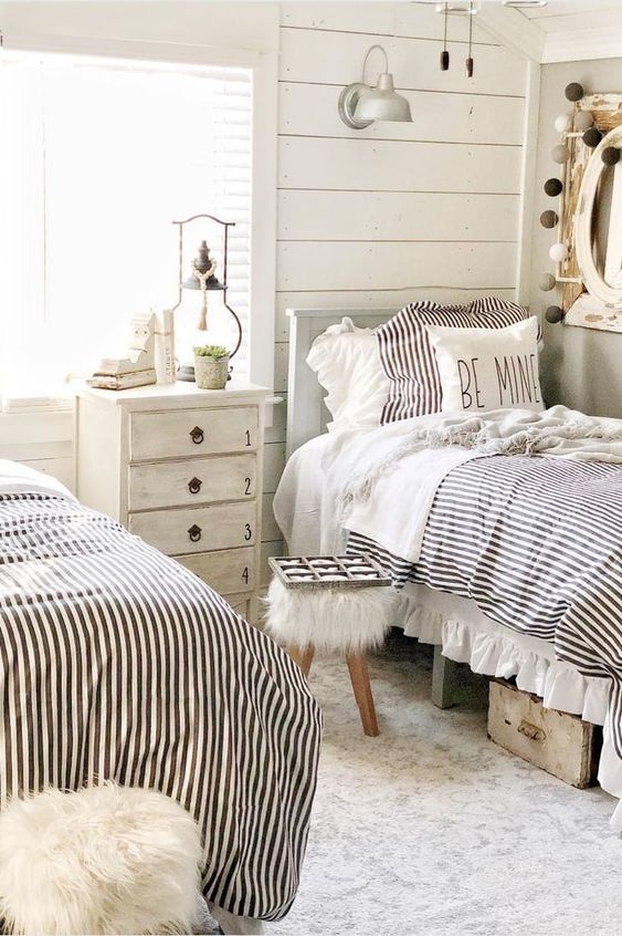 a shared farmhouse kids’ room with whitewashed wooden walls, grey beds, a dresser and a stool and sconces and lamps