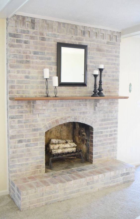 a simple and stylish whitewashed brick fireplace with firewood, a mantel, candles and a mirror for adding coziness to your space