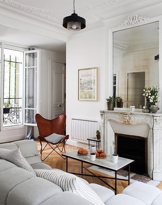 a small yet chic French style living room with an antique fireplace, a coffee table, a brown butterfly chair and a grey sofa