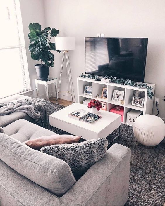 a small yet cozy living room with a small grey sofa, a TV unit, a white table and some lamps plus a potted plant