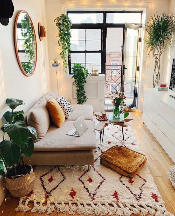 a tiny boho living room with a small neutral sofa, a white storage unit, printed rug, lights and potted greenery