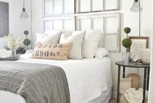 a vintage farmhouse bedroom with a whitewashed headboard composed of window frames, black nightstands, potted plants and pendant lamps