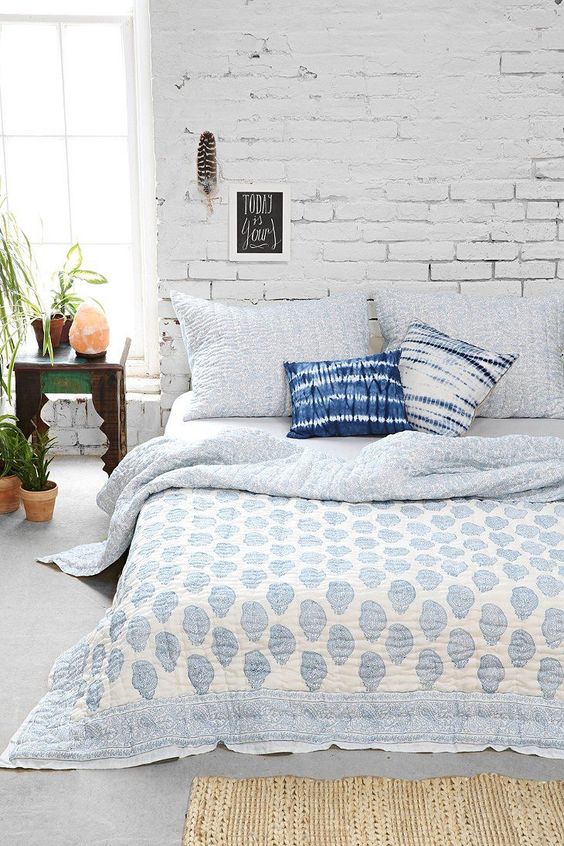 a welcoming light-filled bedroom with a whitewashed brick wall, a bed with catchy bedding, potted plants and some decor