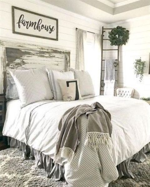 a white farmhouse bedroom with white planked walls, a whitewashed and sahbby chic headboard, neutral bedding, a ladder with a greenery wreath