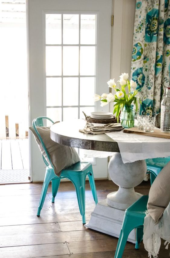 an eclectic dining space with a round table, turquoise metal chairs and blooms plus neutral and turquoise printed curtains