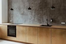 an industrial kitchen with a concrete wall, plywood cabients and a whitewashed floor plus black lamps