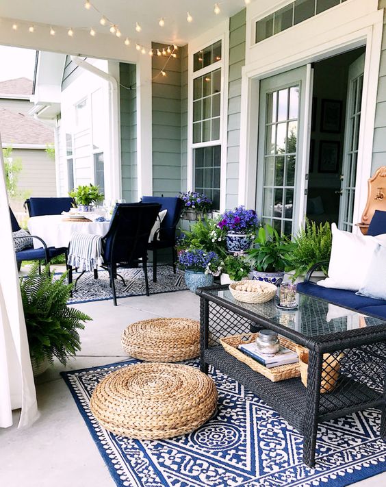 a bold boho summer porch with navy furniture, a printed rug, jute ottomans, a glass coffee table and lots of potted greenery
