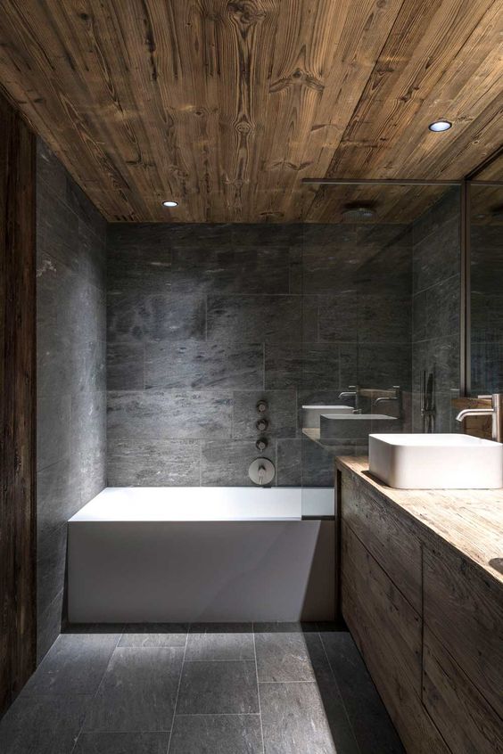 a chalet bathroom clad with grey stone tiles, done with beautiful wood, white appliances and a large mirror is cool