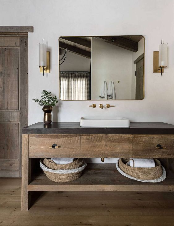 a chalet bathroom done with wood, with a large wooden vanity with a stone countertop, a chic mirror and elegant sconces