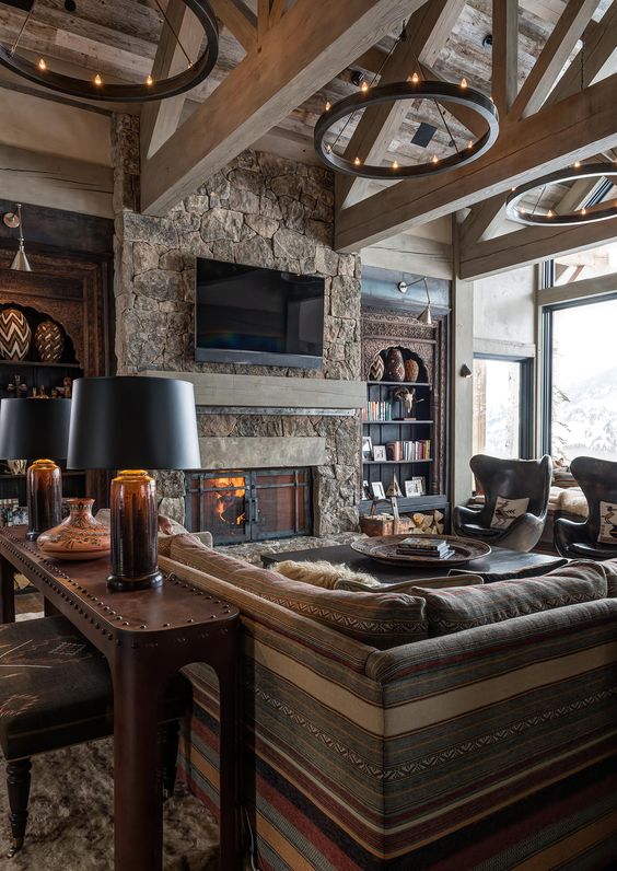 a chalet living room with a fireplace clad with stone, striped seating furniture, metal chandeliers and table lamps