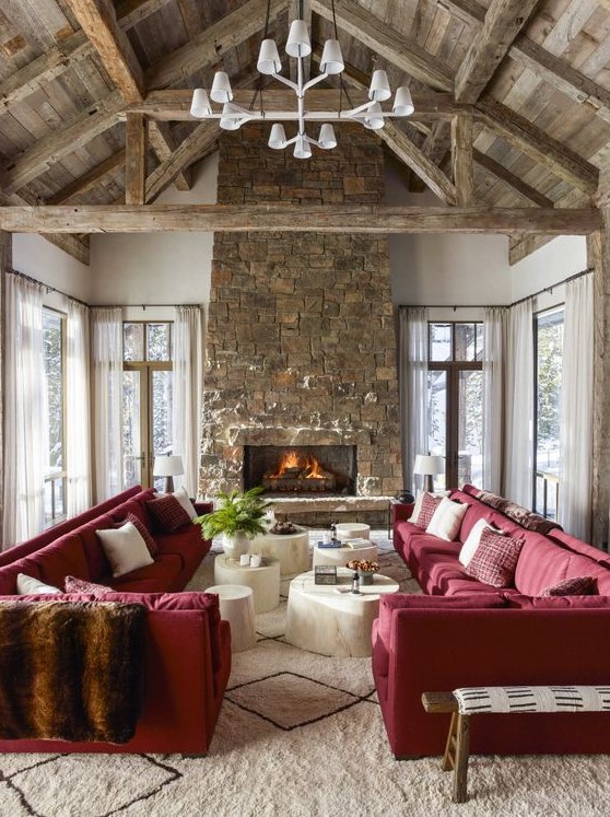 a chalet living room with a wooden ceiling with beams, a stone fireplace, red sofas and whitewashed coffee tables