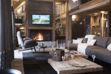 a chalet living room with a wooden ceiling with beams, a wooden coffee table, taupe and blue seating furniture, lights and a fireplace
