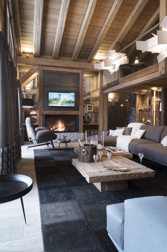 a chalet living room with a wooden ceiling with beams, a wooden coffee table, taupe and blue seating furniture, lights and a fireplace