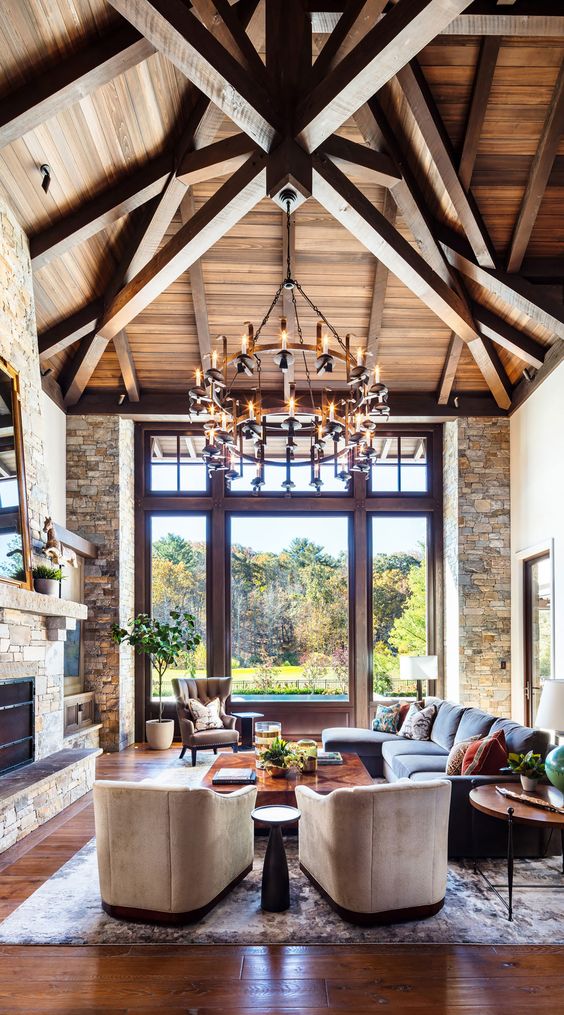a chalet living room with stone walls, a wooden ceiling, wooden beams, a stone clad fireplace, neutral chairs and muted color seating furniture