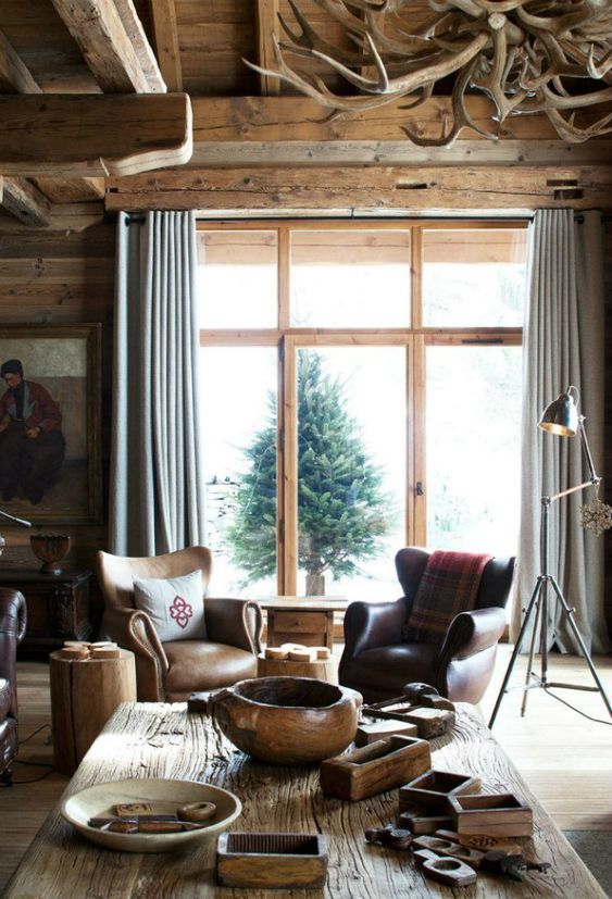a chalet living room with wooden walls and a ceiling, a rough wood coffee table, leather chairs and an antler chandelier