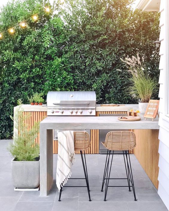 a chic contemporary bbq area of wood and concrete, with a grill and a cooking zone, a concrete dining area and rattan stools