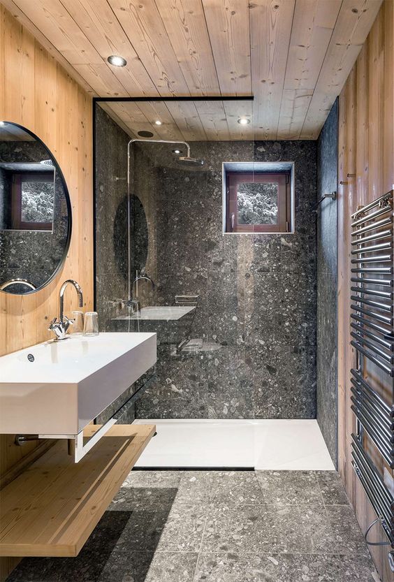 a contemporary chalet bathroom clad with grey terrazzo tiles and light-stained wood, with a shower with a window, a floating vanity
