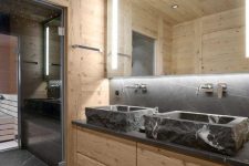 a contemporary chalet bathroom clad with light-stained wood, with stone sinks and a countertop, a lit up mirror and a steam room