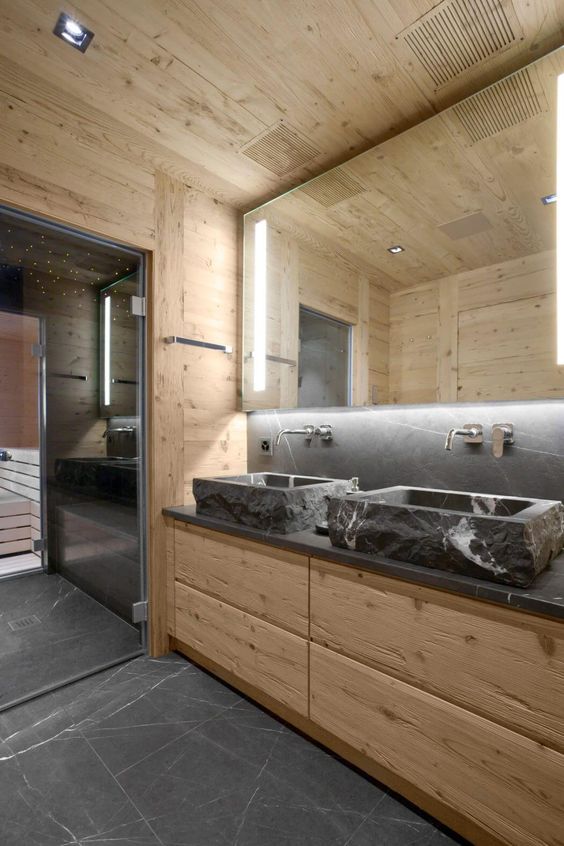 a contemporary chalet bathroom clad with light-stained wood, with stone sinks and a countertop, a lit up mirror and a steam room