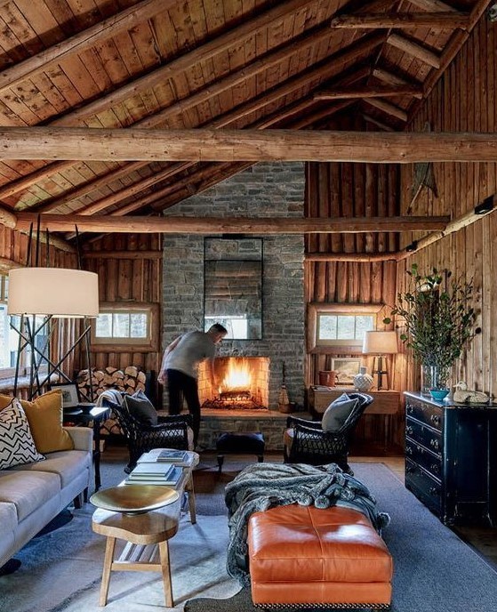 a cozy chalet living room with all wood around, a stone fireplace, a leather ottoman, firewood storage and chic furniture