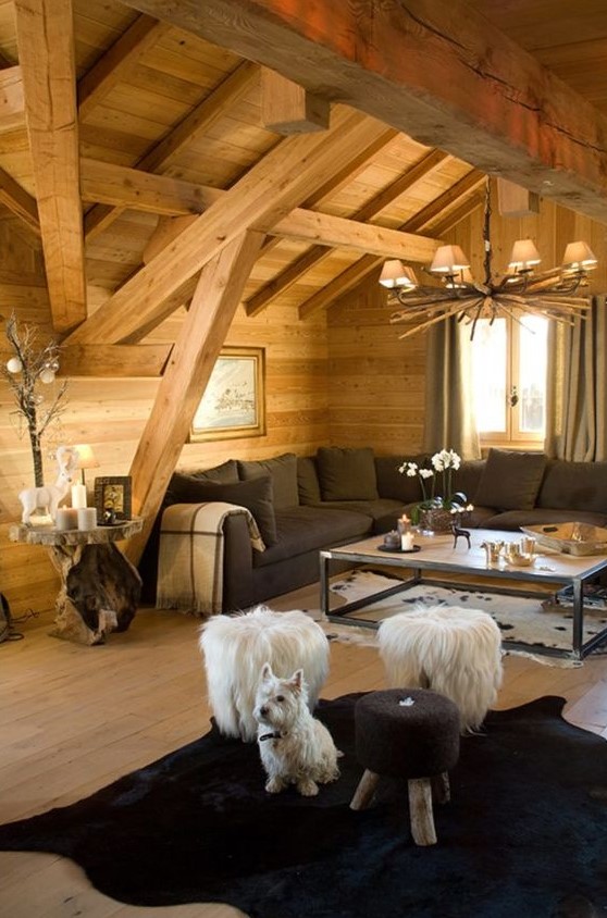 a cozy chalet living room with wooden walls and a ceiling, a rustic chandelier, a black sectional, a black rug, fluffy stools