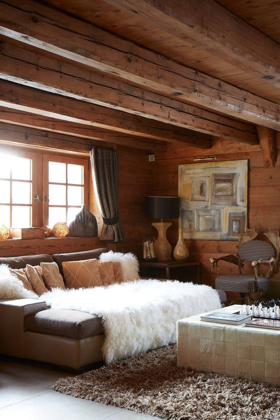 a cozy chalet living space with wooden walls and beams, a grey sofa, a white tile clad table, some art and table lamps