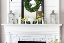 a farmhouse mantel with candle lanterns, vases with bright blooms, a greenery wreath and a mirror