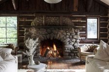 a lovely chalet living room with stained wood walls, a stone fireplace, neutral sofas and round tables, a sphere pendant lamp
