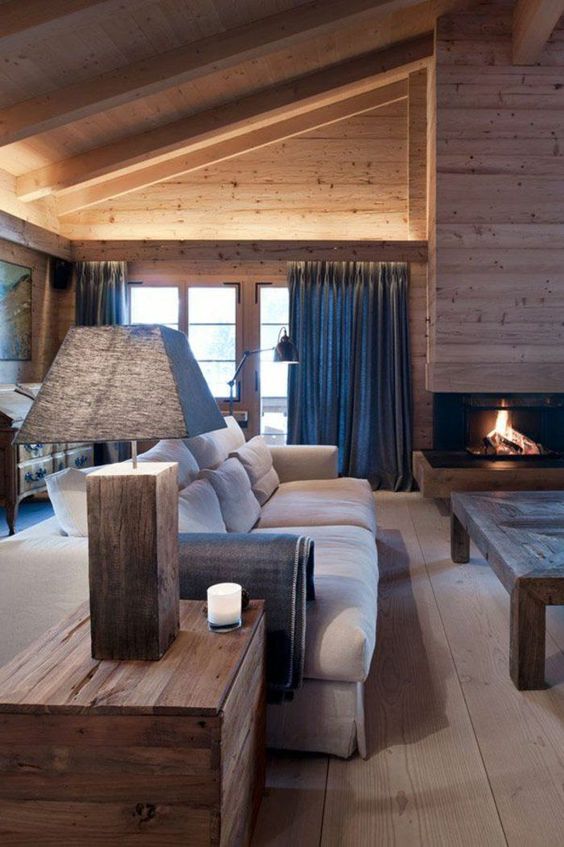 a minimal chalet living room with wooden walls and a ceiling, a fireplace, a neutral sofa, a stained side table and table lamps