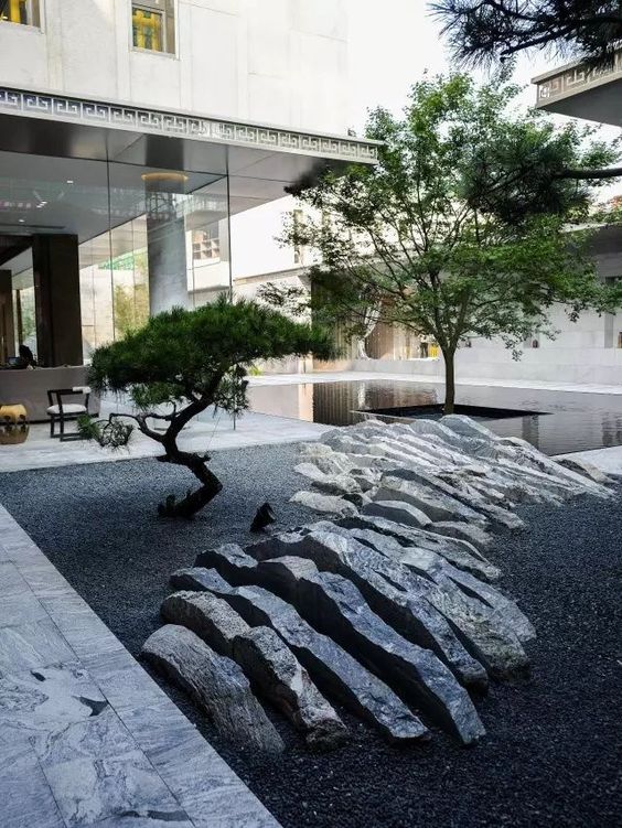 a minimalist Japanese courtyard with a rock that seems to be sliced, two low trees and a sleek pond around one of them