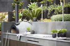 a minimalist outdoor bbq area of wood and concrete, with a grill and a pizza oven plus potted greenery