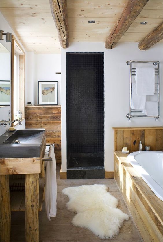 a modern chalet bathroom done with wood, large scale stone tiles, wooden beams on the ceiling, a stone sink and a shower space clad with black tiles