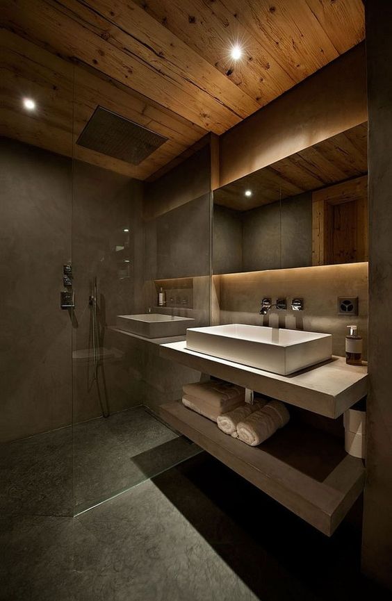 a moody chalet bathroom done with concrete and textural wood, with a floating vanity and a white sink, a lit up mirror