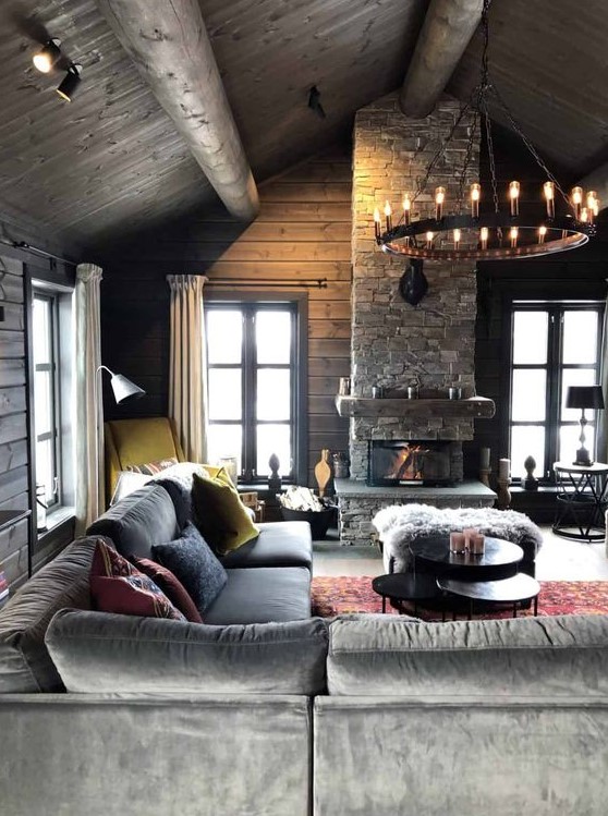 a moody chalet living room with wooden walls and a ceiling, a stone fireplace, a chandelier and comfortable and stylish furniture