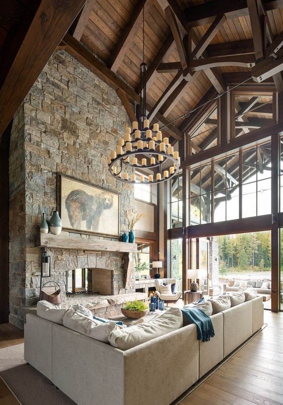 a mountain cabin living room with a wooden ceiling, a stone clad fireplace and stylish neutral furniture plus a statement chandelier