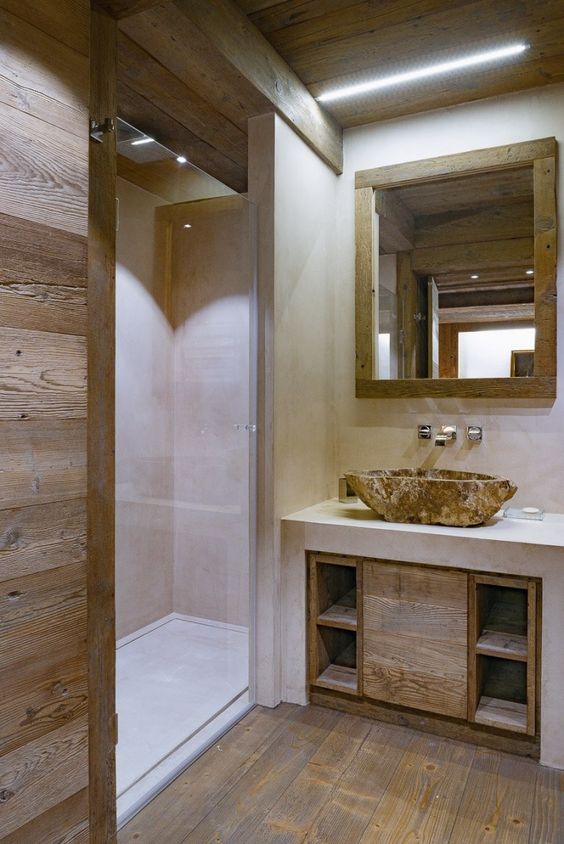 a neutral and contemporary chalet bathroom done with wood and with white concrete, with a stone sink and a mirror in a wooden frame